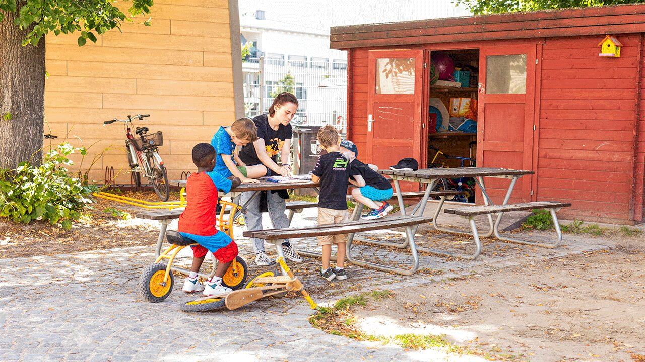 Fachkraft mit Kindern am Spieltisch vor einer Holzhütte